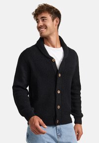 Cárdigan negro de punto con cuello chal, botones de madera y puños acanalados. Tejido texturizado con un ajuste relajado, combinado con una camisa blanca.