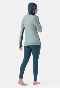 Felpa a maniche lunghe verde chiaro con cappuccio verde scuro, con cuciture arancioni e vestibilità rilassata; abbinata a leggings verde scuro.