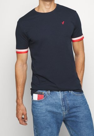 Homme portant un t-shirt bleu marine avec des poignets de manches rayés rouge et blanc, et un jean avec un détail de poche rouge, blanc et noir.