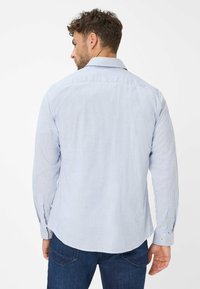 Man met kort krullend haar, gekleed in een lichtblauw en wit gestreept overhemd met lange mouwen en een donkerblauwe spijkerbroek, met zijn rug naar de kijker gekeerd.
