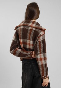 Donna con lunghi capelli castani che indossa una giacca a quadri marrone, arancione e bianca e jeans neri a vita alta, vista da dietro.
