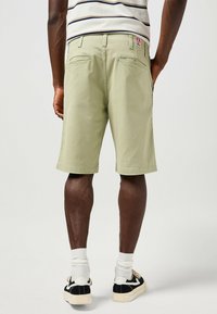 Pantaloni corti verde chiaro in cotone, con una vestibilità classica, due tasche posteriori e un'etichetta con logo cucita. Indossati con calzini bianchi e scarpe da ginnastica nere.