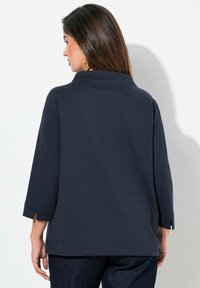 Top blu scuro navy con maniche a tre quarti, un motivo testurizzato e spacchi laterali all'orlo, visto da dietro.