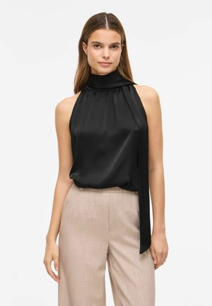 Femme aux longs cheveux bruns portant un chemisier en satin noir sans manches à col montant et un pantalon large beige sur un fond uni.