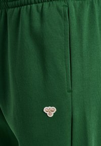 Grønne stofbukser med elastisk linning og et lille beige broderet bi-logo nær lommesømmen på højre side.