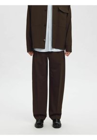 Brune bukser med løs passform, kombinert med en lys blå stripete skjorte og en mørk brun oversized jakke. Sorte sko fullfører antrekket.