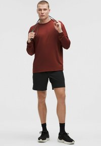 Mans långärmad skjorta i mörkröd tyg, i kombination med svarta shorts. Skjortan har en rund hals och en avslappnad passform. Svarta sneakers fulländar looken.