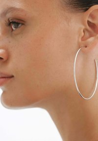 Pendiente grande de aro plateado con un acabado suave y brillante. El diseño presenta una forma circular delgada, usado en la oreja izquierda del modelo.