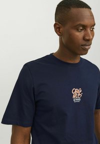 Marineblå bomulds T-shirt med korte ærmer, rund hals og et farverigt grafisk logo trykt på brystet. Glat, let tekstur.