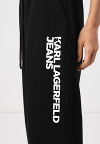 Crne trenirke izrađene od mješavine pamuka s ravnim krojem. Imaju vidljivi bijeli logo "KARL LAGERFELD JEANS" uz bočnu stranu.