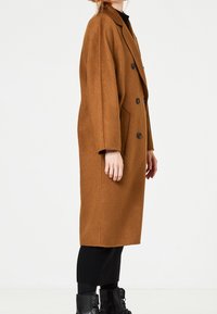 Cappotto lungo in misto lana marrone con design doppiopetto, bottoni neri, tasche laterali e una vestibilità rilassata. Pantaloni neri e stivali in vista.