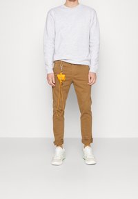 Sweat-shirt gris, pantalon marron fuselé avec revers retroussés, baskets blanches avec des accents verts, et un petit sac jaune attaché par une sangle.