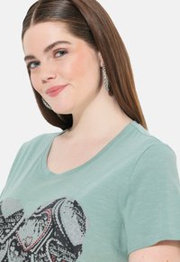 Mintgrøn t-shirt med et hjertegrafik og tekst samt små røde detaljer. Toppen har en v-hals og en blød, let tekstur.