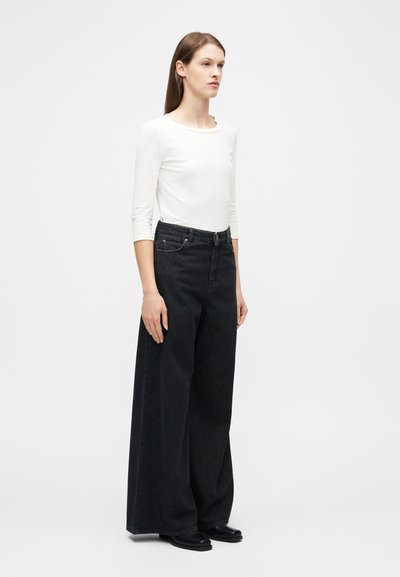 Γυναίκα όρθια, ντυμένη με λευκό πουκάμισο μακριάς μανίκας, μαύρο τζιν παντελόνι με ψηλή μέση και φαρδιά μπατζάκια, και μαύρα παπούτσια, μπροστά σε απλό φωτεινό φόντο.