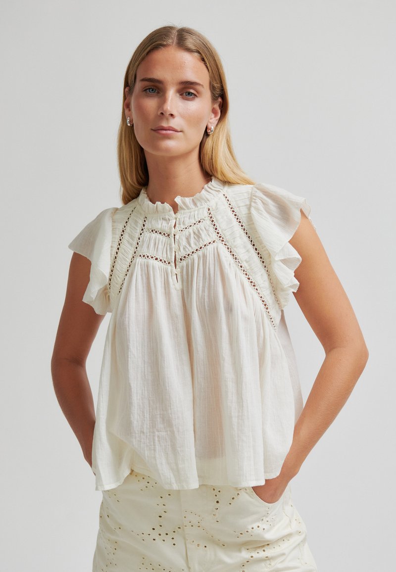 Femme aux longs cheveux blonds portant un chemisier blanc sans manches à bordures à volants et un pantalon blanc assorti en œillet, les mains dans les poches.
