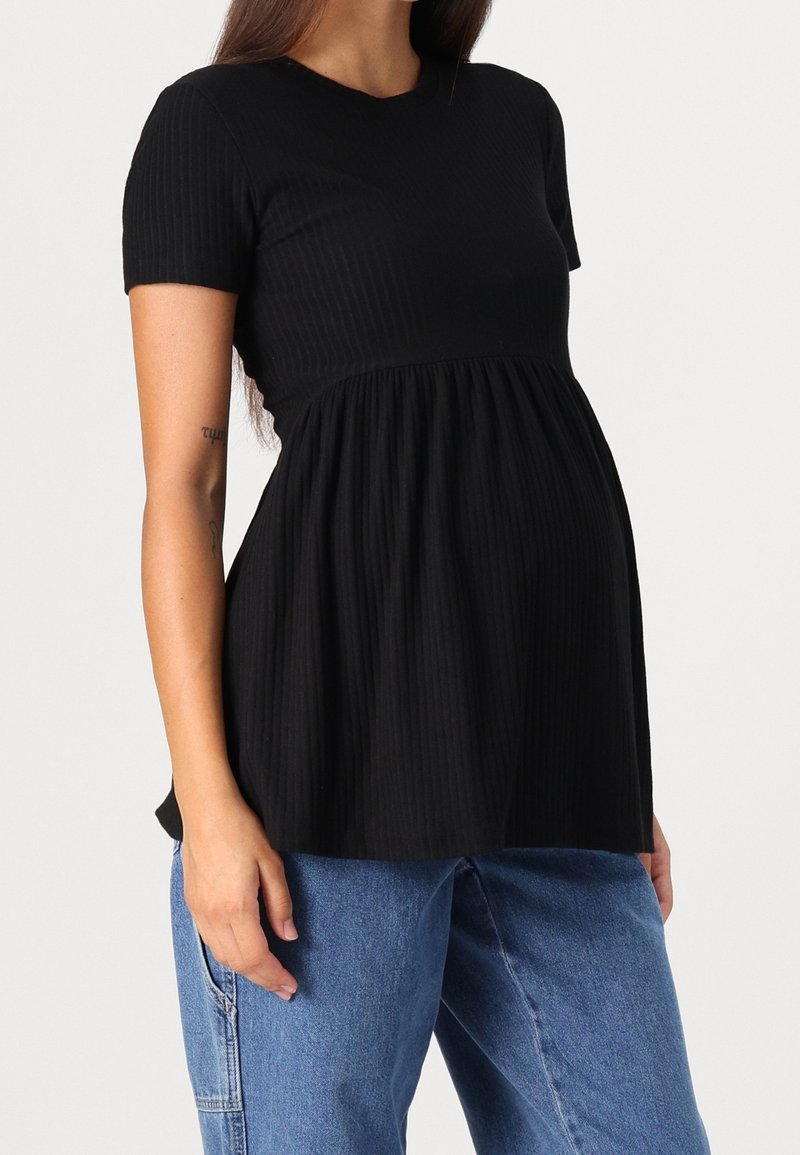 Femme portant un haut de maternité noir à manches courtes avec taille froncée, associé à un jean bleu ample, debout devant un fond uni.
