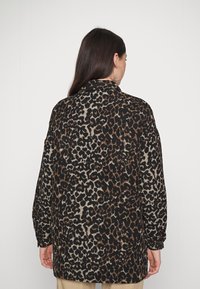 Veste chemise imprimée léopard en tissu doux. Présente des tons noirs, marron et beige, une coupe décontractée et des poignets boutonnés. Vue arrière affichée.