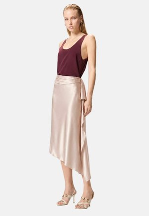 Mujer con top burdeos sin mangas, falda midi asimétrica de satén rosa pálido y sandalias de tacón beige con detalles de cadena.