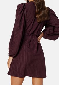 Vestido granate con tela texturizada, mangas largas abullonadas y cintura ajustada. El dobladillo está por encima de la rodilla y presenta sutiles rayas verticales.