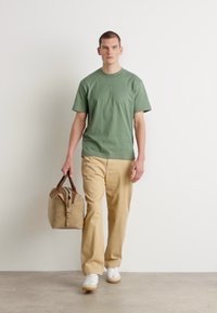 T-shirt en coton vert, coupe décontractée, associée à un pantalon beige. Le modèle porte un sac beige avec un rabat et des anses supérieures. Baskets blanches.