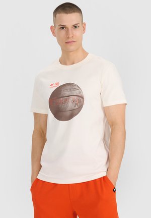 Młody mężczyzna w kremowym T-shircie z brązową grafiką koszykówki i pomarańczowych szortach, stojący z rękami w kieszeniach na jednolitym tle.