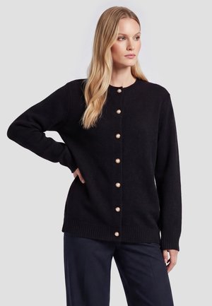 Kvinde med langt blondt hår iført en sort knappet cardigan med perle-lignende knapper og mørke bukser, stående med den ene hånd på hoften.