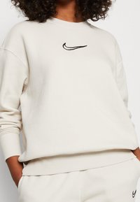 Crèmefärgad sweatshirt i mjukt tyg, med en svart Nike-logga på bröstet, ribbade ärmslut och en avslappnad passform.