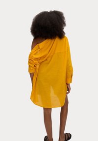 Camicia gialla di dimensioni oversized con maniche arrotolate e design spalla scoperta. Tessuto leggero e testurizzato con bottoni sulla spalla.