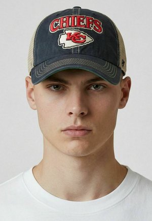 Junger Mann, der eine marineblaue und beige Kansas City Chiefs Baseballkappe und ein weißes T-Shirt mit Rundhalsausschnitt trägt, blickt mit neutralem Gesichtsausdruck nach vorne.