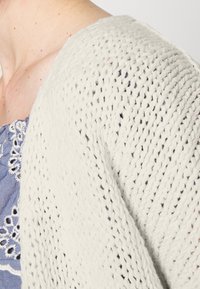 Helle cremefarbene Strickjacke mit offenem Gewebe und lockerer Passform, getragen über einem blauen bestickten Oberteil mit weißen Details.