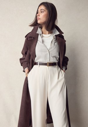 Žena s kratkom tamnom kosom, odjevena u smeđi baloner, prugastu košulju preko bijelog topa, krem hlače i zlatne dodatke, ruke u džepovima.