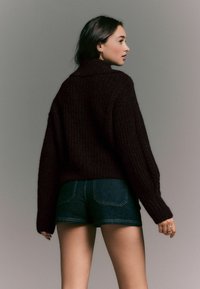 Brauner gerippter Strickpullover mit hohem Kragen, kombiniert mit dunklen Jeansshorts. Das Model steht leicht seitlich, was den strukturierten Stoff betont.