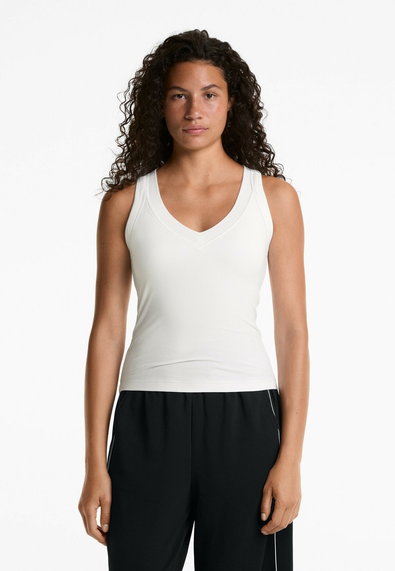 Débardeur blanc côtelé avec un col en V, design ajusté et larges bretelles, associé à un pantalon noir avec des accents blancs.