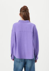 Relaxed-fit, long-sleeve shirt in light purple, made of textured fabric, featuring a fold-down collar and buttoned cuffs. Paired with blue denim jeans.
