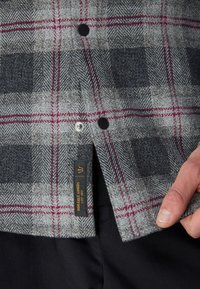 Cămașă cu carouri gri, dintr-un amestec de lână, cu nasturi negri și accente roșii, având un etichetă de marcă vizibilă aproape de margine.