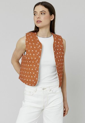 Gilet matelassé en orange rouille avec broderie florale. Présente un col rond et un design sans manches, porté par-dessus un haut en maille blanche.