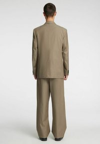 Bež oplet s širokimi hlačnicami in strukturiranim blejzerjem, ki ima dva gumba in gladko teksturo. Enostaven pogled od zadaj.