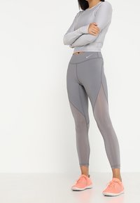 Vrouw in een lichtgrijze lange mouwtop en grijze Nike leggings met gaaspanelen, staand met gekruiste armen en met heldere koraalkleurige sportschoenen.