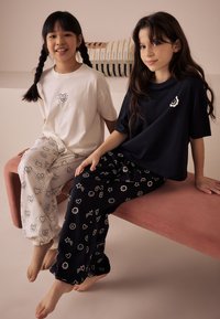 Deux enfants portent des ensembles de pyjamas assortis. L'un a un t-shirt blanc avec un motif en forme de cœur, l'autre un t-shirt noir avec des motifs floraux. Tissu doux, coupe décontractée.