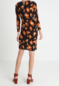 Robe noire avec motif abstrait orange, manches longues, détail dos en trou de serrure, forme ajustée, associée à des sandales à talons blocs strappées marron.
