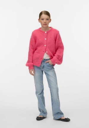 Junge Person steht mit einer Hand in der Tasche, trägt eine leuchtend pinke, geknöpfte Strickjacke, hellblaue Jeans und schwarze Ballerinas.