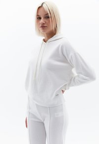 Sudadera blanca con capucha, cordones y textura acanalada, combinada con pantalones ajustados blancos a juego que presentan detalles de costura y un pequeño logo.