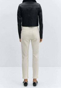 Chaqueta de cuero negra corta con cuello alto, combinada con pantalones ajustados de color beige claro. Zapatos negros con hebillas decorativas.