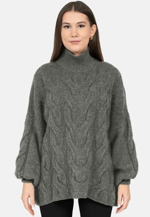 Junge Frau trägt einen grauen, grobgestrickten Rollkragenpullover mit Ballonärmeln und steht vor einem einfachen hellen Hintergrund.