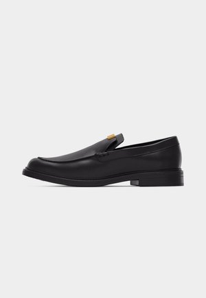 Schwarzer Leder-Slipper mit rundem Zehenbereich, niedrigem Absatz und goldenem Akzent am Obermaterial vor schlichtem weißem Hintergrund.