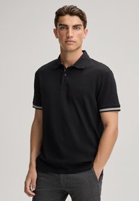 Zwarte polo met een textuurstof, korte mouwen met gestreepte manchetten en een subtiel logo op de borst. Gecombineerd met grijze broek.