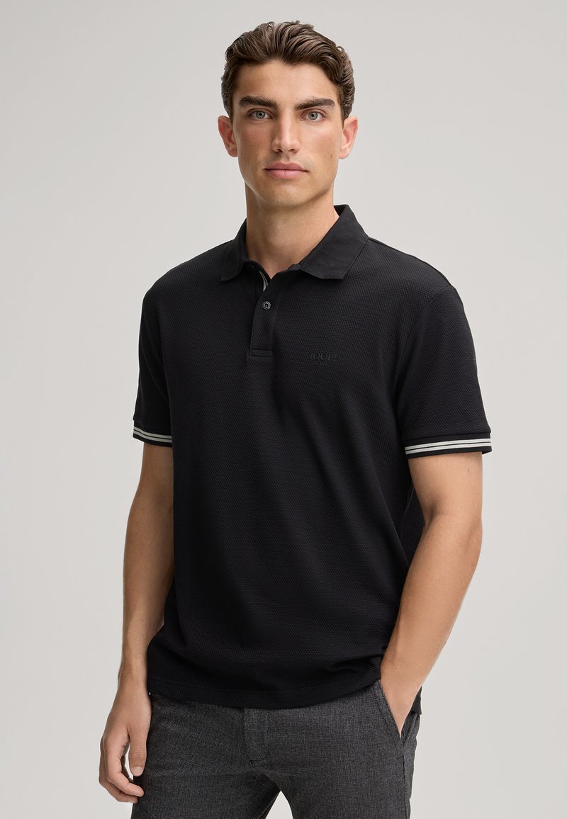 Zwarte polo met een textuurstof, korte mouwen met gestreepte manchetten en een subtiel logo op de borst. Gecombineerd met grijze broek.