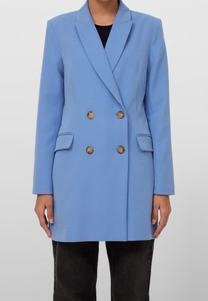 Vrouw draagt een lichtblauwe dubbelbreasted blazer met bruine knopen en klepzakken, gecombineerd met een zwarte broek.