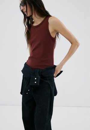 Femme portant un haut sans manches bordeaux avec bordure en dentelle, pantalon noir, et une veste sombre nouée autour de la taille, debout devant un fond blanc.