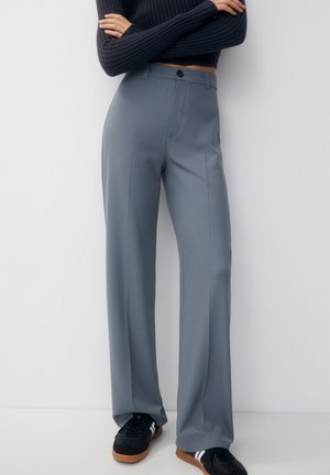 Personne portant un pantalon gris taille haute sur mesure, un pull court noir à côtes, et des baskets noires à rayures blanches, debout contre un mur blanc.